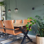 lichte vloer in houtpatroon met brede planken in licht eiken, stijlvolle eettafel met zwart metalen onderstel en tafelblad in blank hout, omringd door comfortabele cognackleurige leren stoelen met stalen frames, tegen een groene muur met drie hanglampen met glazen kap en groene planten in rieten manden