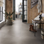 Antracietgrijze tegelvloer met grote, effen tegels in een matte, gladde structuur in een moderne hal met houten wandbetimmering en metalen meubelen.