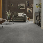 grijze tegelvloer met grote, matte tegels in licht antraciet met subtiel golvend streeppatroon, moderne woonkamer met donkergroene fluwelen fauteuil, zwarte staande lamp, ronde houten salontafel en planten op de vloer