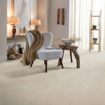 Licht beige tegelvloer met grote matte tegels en subtiel fijnkorrelig oppervlak, gecombineerd met een lichtgrijze fauteuil met donker houten poten en een grof gebreide taupe deken.