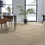 lichte vloer in visgraatpatroon met brede planken, licht beige eiken kleur en een subtiele houtnerfstructuur, gecombineerd met bruine gestoffeerde eetkamerstoelen en een zwarte eettafel in een lichte, moderne kamer met grote raampartijen en groene planten in zwarte potten