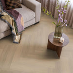 lichte beige eiken vloer met visgraat patroon, matte afwerking, lichte houtnerfstructuur zichtbaar, gecombineerd met een beige bank en een houten bijzettafel met vaas en bloemen