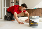 Man in rode shirt die PVC lijm uitsmeert op een lichte ondervloer in een woonkeuken met kastfronten in donkerbruin houtdecor.