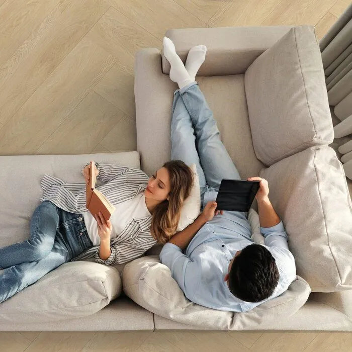 licht eiken visgraatvloer met smalle planken in rustig patroon en zacht matte afwerking, zittend op beige stoffen hoekbank een vrouw leest een boek en een man gebruikt een tablet