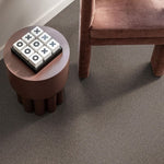 taupe tapijt met fijne, zachte poolstructuur naast een rond, mat bruin tafeltje en een opengewerkt bruin fluwelen fauteuilbeen