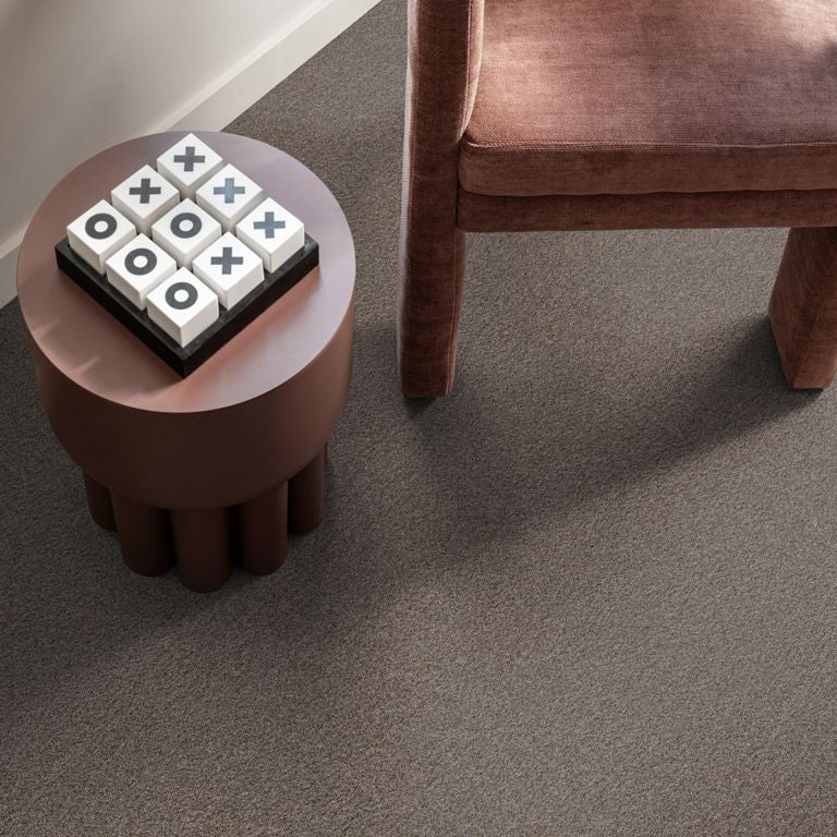 taupe tapijt met fijne, zachte poolstructuur naast een rond, mat bruin tafeltje en een opengewerkt bruin fluwelen fauteuilbeen