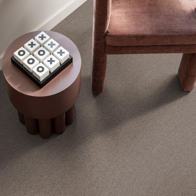Taupe kleurige zachte tapijtvloer met fijne pluizige structuur, met bruine fluwelen stoel en ronde donkerbruine bijzettafel met groot dambordspel.