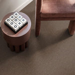 taupe bruin tapijt met fijne structuur en matte afwerking onder een rond bijzettafeltje en een fluweelzachte, roestbruine fauteuilpoot