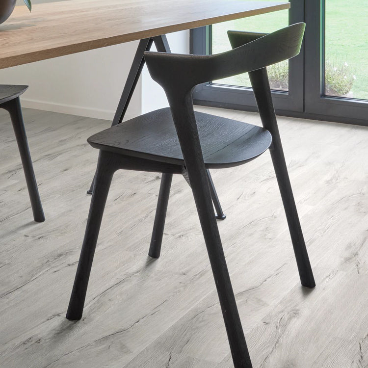 lichte vloer met brede planken in licht grijs eiken houtstructuur, mat afgewerkt, met een moderne zwarte houten stoel en houten tafelpoten in een lichte, minimalistische kamer naast groot raam