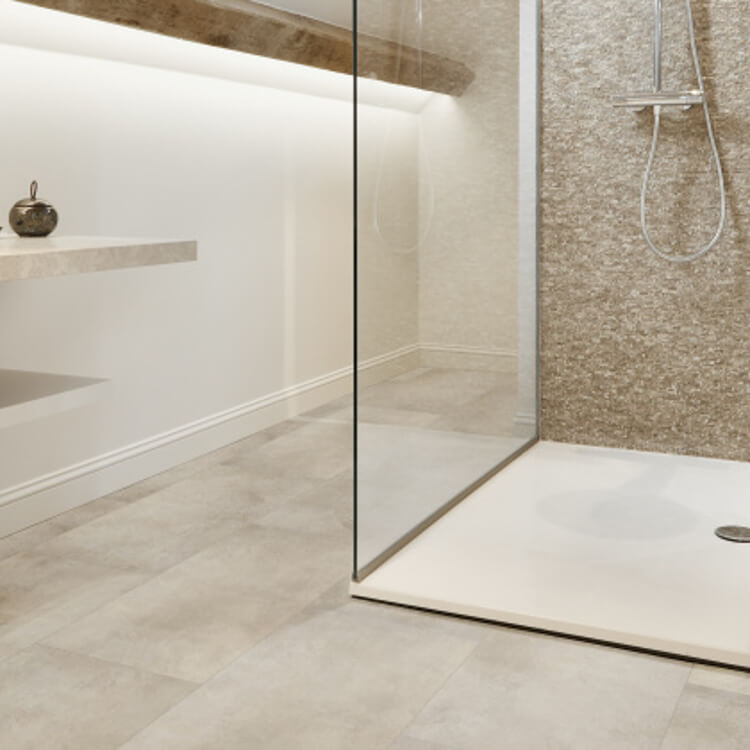 Tegelvloer met grote vierkante licht taupe tegels met een subtiel golvend streeppatroon en een douchegedeelte met een lichte beige natuurstenen wandafwerking in een fijnkorrelige structuur.