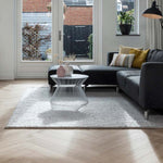 Lichte eiken visgraatvloer met subtiele houtstructuur in woonkamer, grijs vloerkleed met grof geweven structuur, donkergrijze hoekbank met diverse kussens in mosterdgeel en zacht beige, witte moderne bijzettafel van draadmetaal.