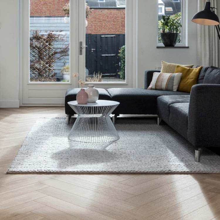 Lichte eiken visgraatvloer met subtiele houtstructuur in woonkamer, grijs vloerkleed met grof geweven structuur, donkergrijze hoekbank met diverse kussens in mosterdgeel en zacht beige, witte moderne bijzettafel van draadmetaal.