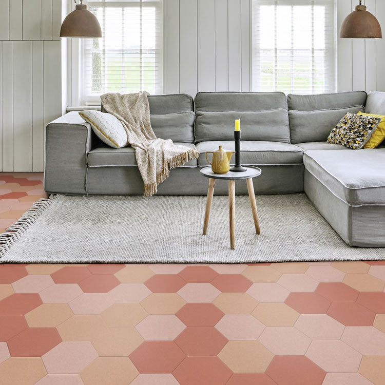 Hexagonale tegelvloer in zachte, warme tinten met terracotta, licht beige en zandkleurige zeshoekige tegels, gecombineerd met een lichtgrijs wollen vloerkleed, grijze stoffen hoekbank en lichte inrichting met witte houten wanden.