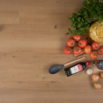 Licht eiken vloer met brede planken en subtiele houtnerfstructuur, als achtergrond voor keukeningrediënten zoals tomaten, kruiden, knoflook, uien, pasta en een fles olijfolie.