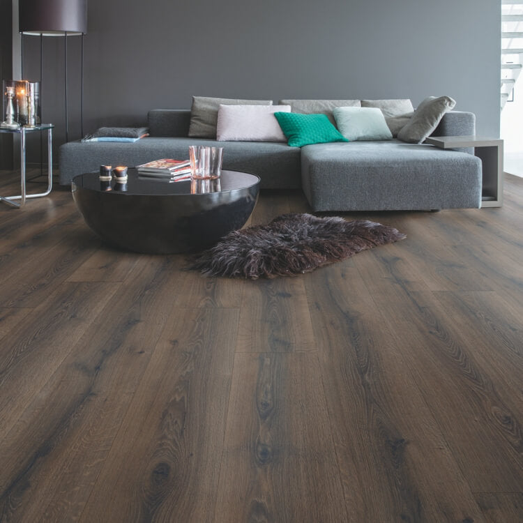 Donker bruin eiken vloer met brede planken en natuurlijke houtstructuur in moderne woonkamer met grijze hoekbank en zwarte ronde salontafel.