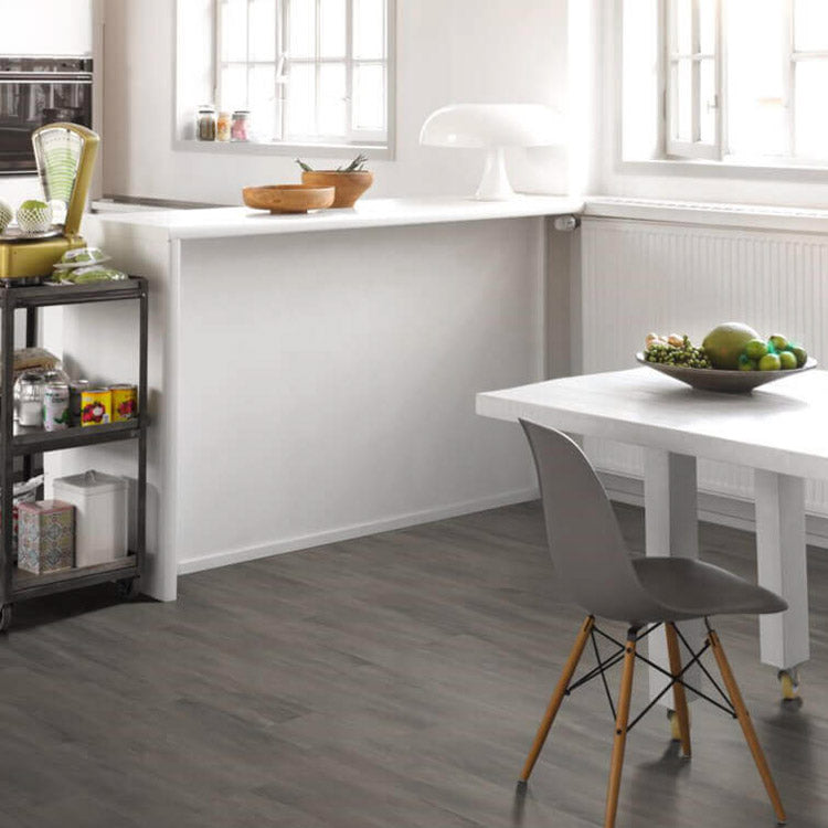 Donkergrijze vloer met brede planken in een houtstructuur, gecombineerd met een witte eettafel en grijze kunststof stoel met houten poten in een lichte, moderne keukenruimte.