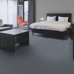 donkergrijze tegelvloer met fijne, subtiele korrelstructuur in moderne hotelkamer met zwart bedframe, wit beddengoed, oranje fauteuil en donker houten salontafel