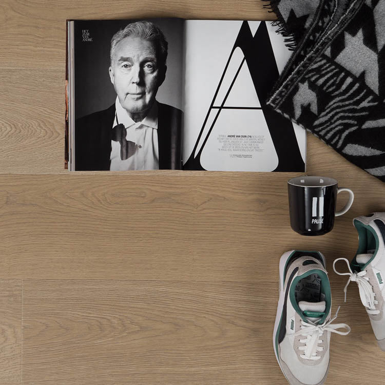 lichte laminaatvloer in eikenstructuur met fijne houtnerf en subtiele beige tint, met opengevouwen zwart-wit magazine, zwart-witte geprint deken, zwarte mok met witte tekst en wit-groene sneakers op de vloer geplaatst