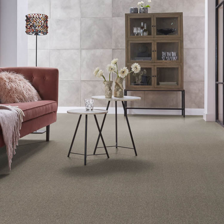 Taupe kleurig fijngeweven tapijt met subtiele structuur in een moderne woonkamer met salontafels en kast tegen een betonnen wand.