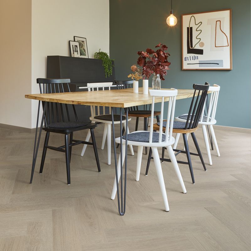 Licht eiken vloer met visgraatpatroon en brede planken, moderne eetkamer met zwarte en witte stoelen aan houten tafel, minimalistische inrichting met groene muur en abstract schilderij.
