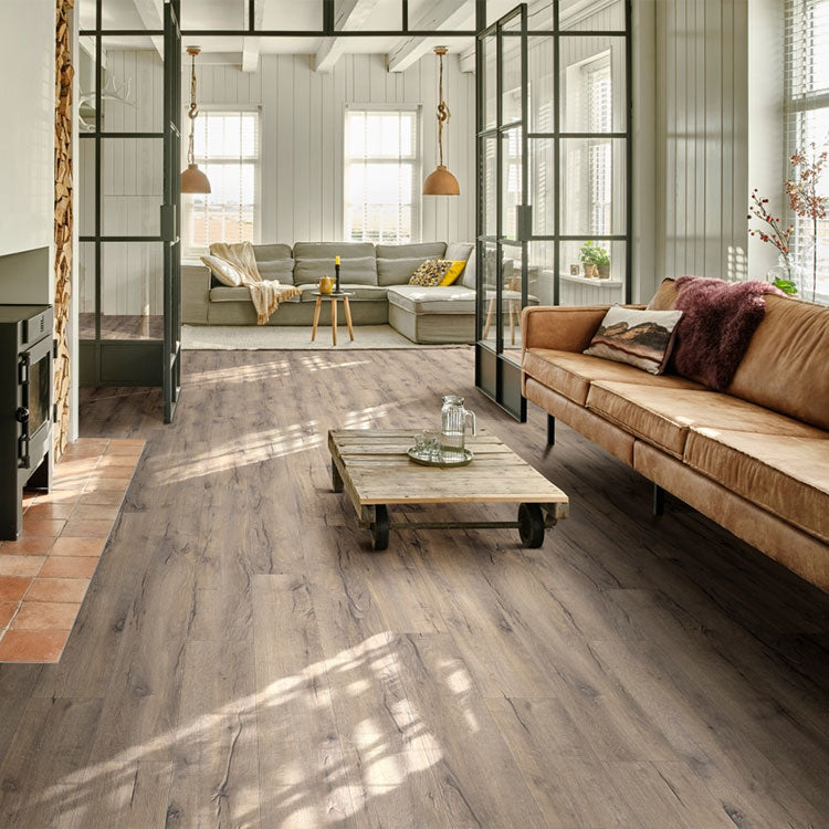 Ruime woonkamer met vloer in licht grijs bruin eiken kleur met brede planken en subtiele houtnerven, gecombineerd met een cognackleurige leren bank en een robuuste houten salontafel op wielen.