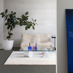 donkergrijze vlakke tegelvloer met een lichte, zachte witte hoogpolige tapijttegel, een beige viscose bank, glazen salontafel met witte geometrische poten en paarse en blauwe kaarshouders, grote groene kamerplant in witte pot tegen een lichtgrijze muur
