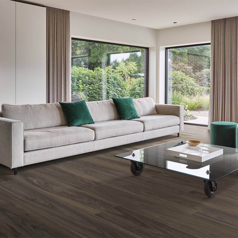 donkergrijze houten vloer met lange brede planken en een natuurlijke, licht gegolfde houtstructuur in een moderne woonkamer met beige bank en groene kussens, glazen salontafel op wielen en grote ramen met zicht op tuin