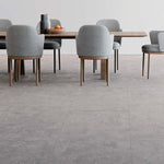 Tegelvloer met grote tegels in licht grijs betonstructuur, moderne eetkamer met grijze gestoffeerde stoelen en houten tafelpoten.