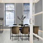 Lichte grijs eiken vloer met brede planken in een moderne eetkamer met stijlvolle donkere stoelen, een houten tafel en een vaas met takken, rondom drie hoge ramen met smalle jaloezieën.