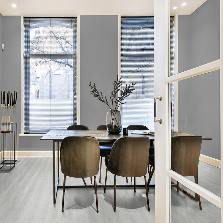Lichte grijs eiken vloer met brede planken in een moderne eetkamer met stijlvolle donkere stoelen, een houten tafel en een vaas met takken, rondom drie hoge ramen met smalle jaloezieën.