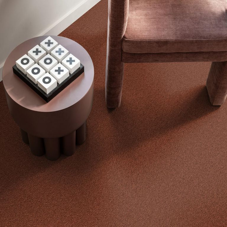 Tapijt in warme roodbruine kleur met fijne, dichte structuur, naast een ronde bruine bijzettafel met spelletje en een stoel met bekleding in een bijpassende roodbruine stof.