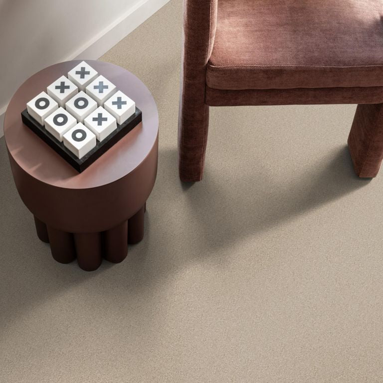 taupe pvc vloer met fijne zandstructuur, naast een ronde donkerbruine bijzettafel met een kruis-en-rondspel van houten blokken en een kastanjebruine gestoffeerde stoel