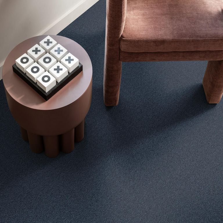 donkerblauw antracietkleurig hoogpolig tapijt met zachte structuur onder bruine stoffen stoel en bruin rond bijzettafeltje met een kruisjes-en-nulletjestegelspel erop