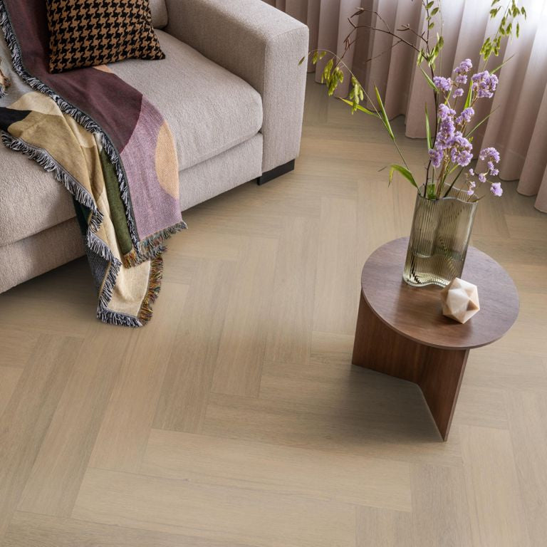 lichte beige eiken vloer met visgraat patroon, matte afwerking, lichte houtnerfstructuur zichtbaar, gecombineerd met een beige bank en een houten bijzettafel met vaas en bloemen