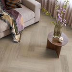 Licht beige eiken visgraatvloer met subtiele houtnerfstructuur in een moderne woonkamer naast een beige bank en een ronde houten salontafel met vaas en bloemen.