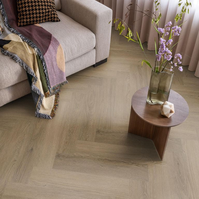 lichte vloer in houtpatroon visgraat van licht beige eiken met subtiele nerfstructuur in een moderne woonkamer naast een beige bank, met een kleine ronde houten tafel en een vaas met paarse bloemen