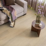 lichte beige eiken vloer met brede planken, subtiel houtnerfstructuur, gecombineerd met een beige stoffen bank en een houten bijzettafel met vaas en paarse bloemen