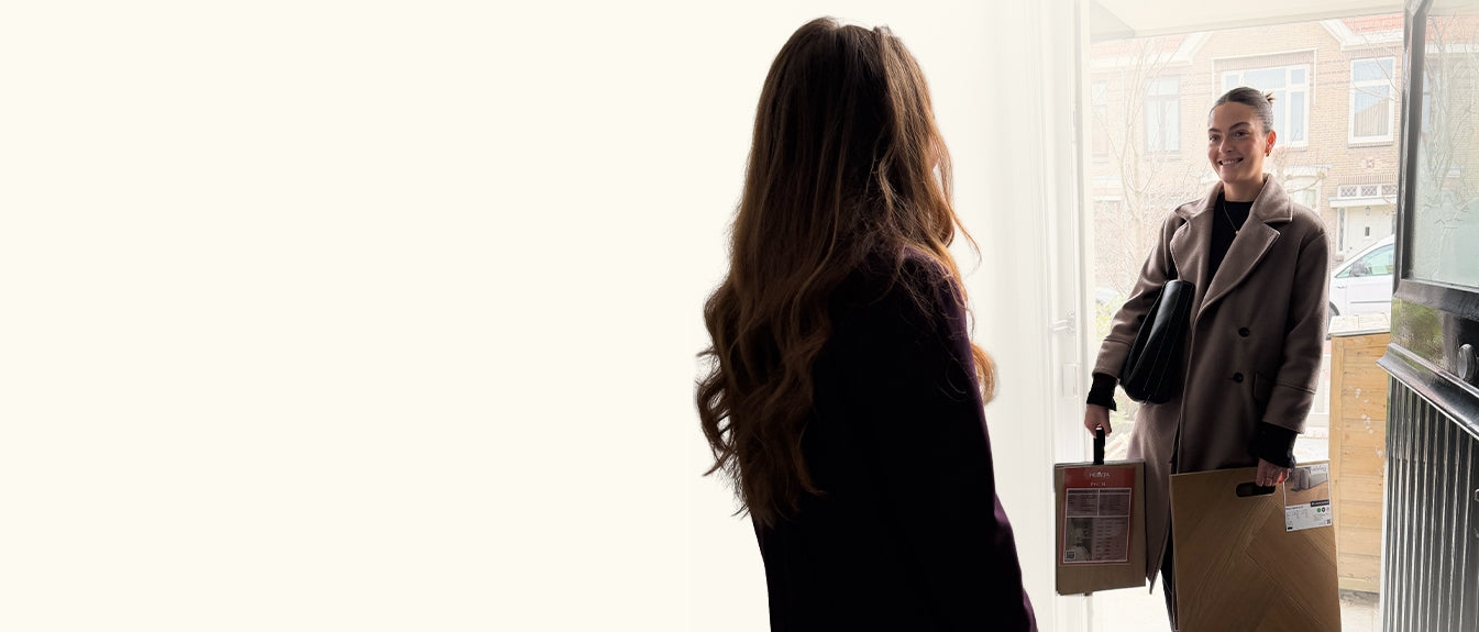 Vrouw bij de voordeur die een licht beige eiken vloer sample en een papieren tas met vloerproducten overhandigt aan een andere vrouw binnen in huis, met natuurlijk licht vanuit de deur.