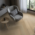 lichte eiken vloer in visgraat patroon met brede planken, grijze stoffen fauteuil en ronde houten bijzettafel naast raam met witte vitrage curtains