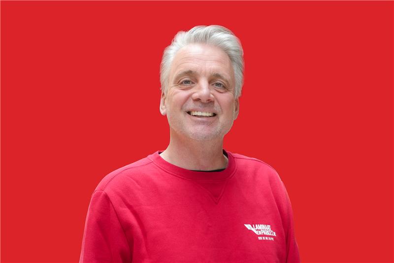 Man met grijs haar in rode trui met wit logo van Laminaatenparket.nl, staand voor een effen rode achtergrond.