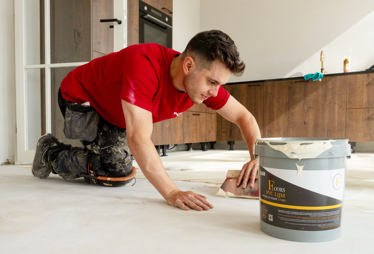 Man in rode shirt die PVC lijm uitsmeert op een lichte ondervloer in een woonkeuken met kastfronten in donkerbruin houtdecor.