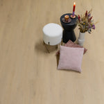 licht beige eiken vloer met fijne, natuurlijke houtnerf en knoestpatroon, brede planken, matte afwerking, gestyled met wit bouclé krukje, zwart bijzettafeltje met multicolor rode naar paars verlopende kaars, kopje koffie op paars schoteltje en vaas met droogboeket naast twee zachte kussens in pastelroze tinten