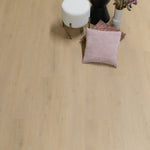 lichte beige eiken vloer met subtiele houtnerfstructuur en brede planken, met een wit gebogen poefje, zwarte vaas en zachte kussens in roze tinten en decoratieve drogende bloemen
