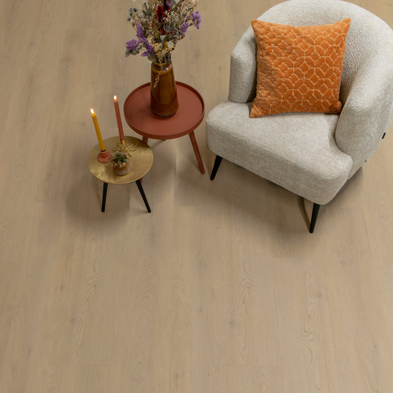 lichte beige eiken vloer met brede planken en een subtiele houtnerfstructuur, gecombineerd met een crème kleurige geborgen fauteuil met oranje velours kussen en twee bijzettafels in aardetinten met decoratieve kaarsen en droogbloemenvas