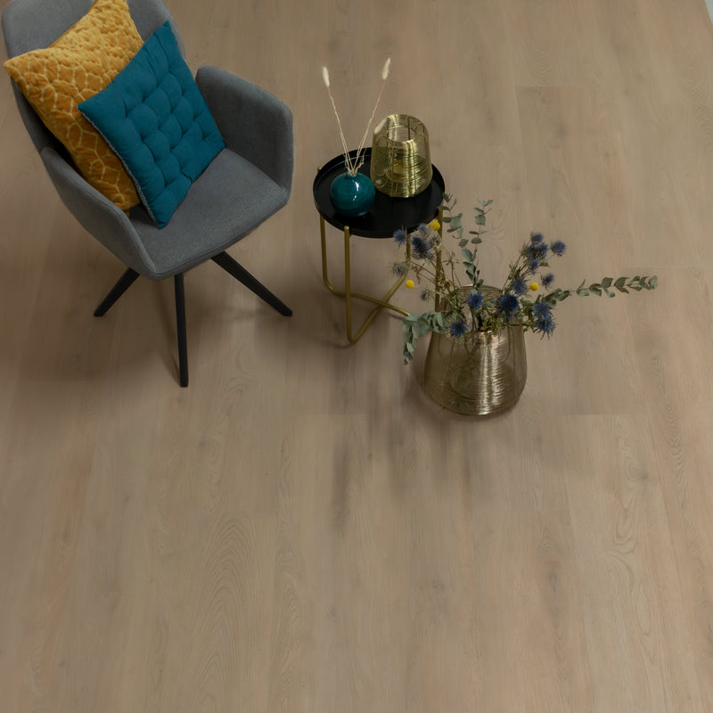 lichte eiken vloer met brede planken en subtiele houtnerfstructuur, grijze fauteuil met blauwe en mosterdgele kussen, zwart bijzettafeltje met gouden pot en vaas met droogbloemen