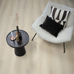 licht eiken vloer met brede planken en subtiele houtnerfstructuur in een rustige, lichte woonkamer met creme stoffen fauteuil en zwart-wit sierkussens naast een zwart metalen bijzettafeltje met decoratieve vaas en keramische kandelaars