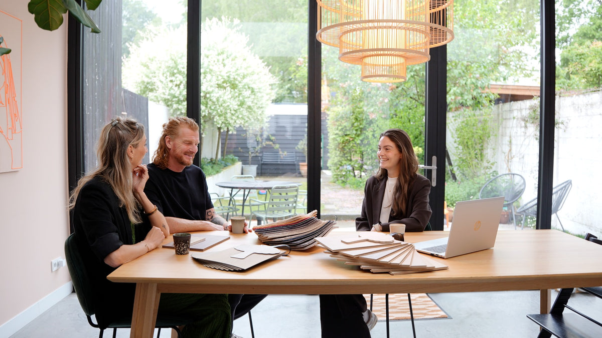 Drie personen in een lichte, moderne vergaderruimte met groot raam naar tuin, houten tafel met stalen poten, verschillende staaltjes vloer- en materialenmonsters in naturel tinten en een grote hanglamp met rotan structuur erboven.