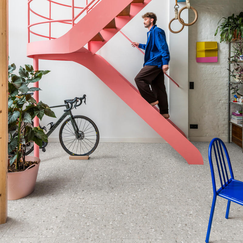 lichtgrijze tegelvloer met groot terrazzo patroon van gemêleerde korrels in verschillende tinten grijs en wit in een moderne woonruimte met een roze metalen trap en diverse accessoires