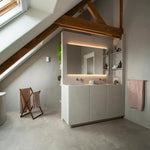 lichte betonnen vloer met een gelijkmatige, zachte structuur en matte afwerking in een moderne badkamer met witte wastafelmeubel en houten balken onder een schuin dak raam