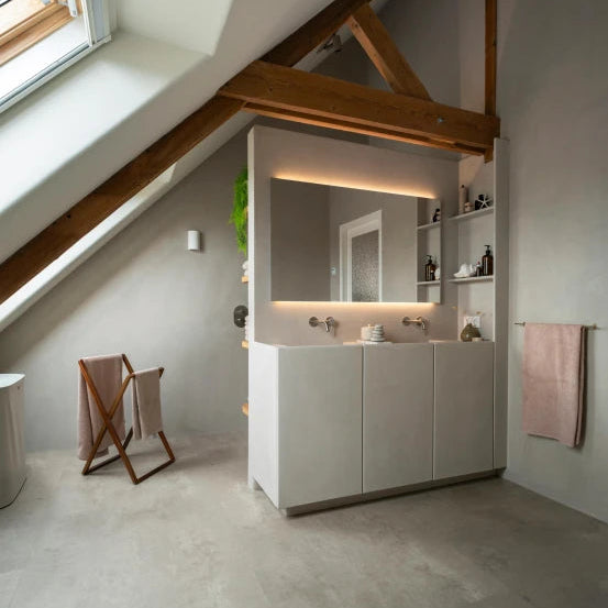lichte betonnen vloer met een gelijkmatige, zachte structuur en matte afwerking in een moderne badkamer met witte wastafelmeubel en houten balken onder een schuin dak raam